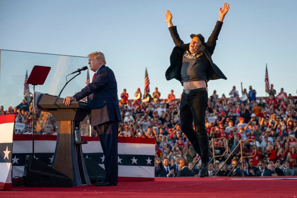 Elon Musk jumps foolishly on stage next to disgraced former president Donald Trump.