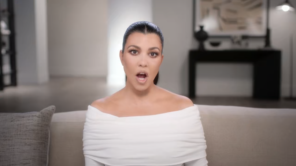 Kourtney Kardashian tells the confessional camera what she's thinking with her mouth open, wearing a wide-necked white blouse.