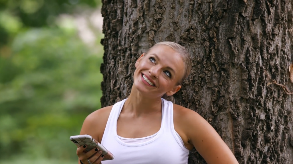 Mackenzie McKee leans against a tree and smiles while wearing a white tank top.